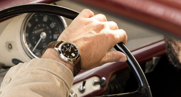 A wrist shot of a watch by Reservoir holding a steering wheel in a classic Porsche